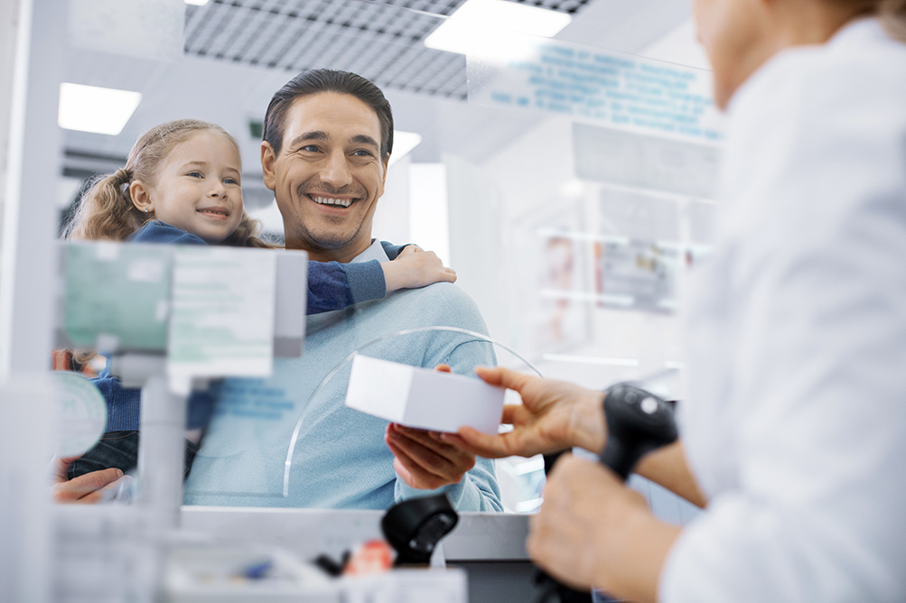 Caucasian male holding a caucasian girl while picking up a prescription at the pharmacy counter.